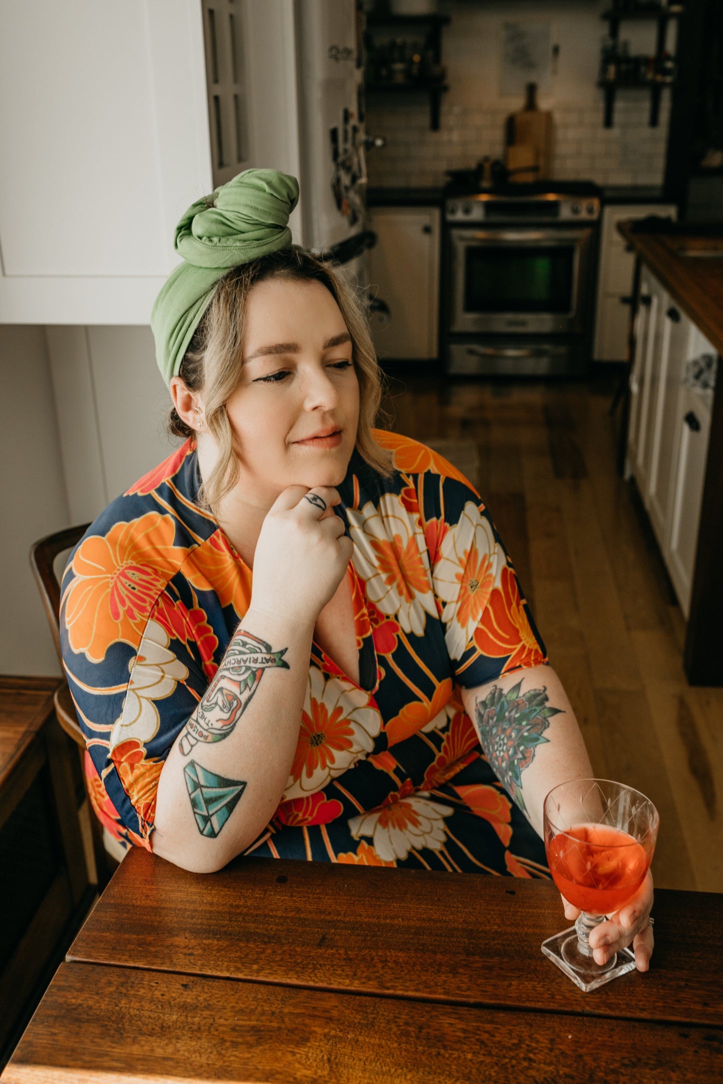 woman sits down for a drink in the kitchen while quickly drying her hair with  her green Capillum bamboo hair towel